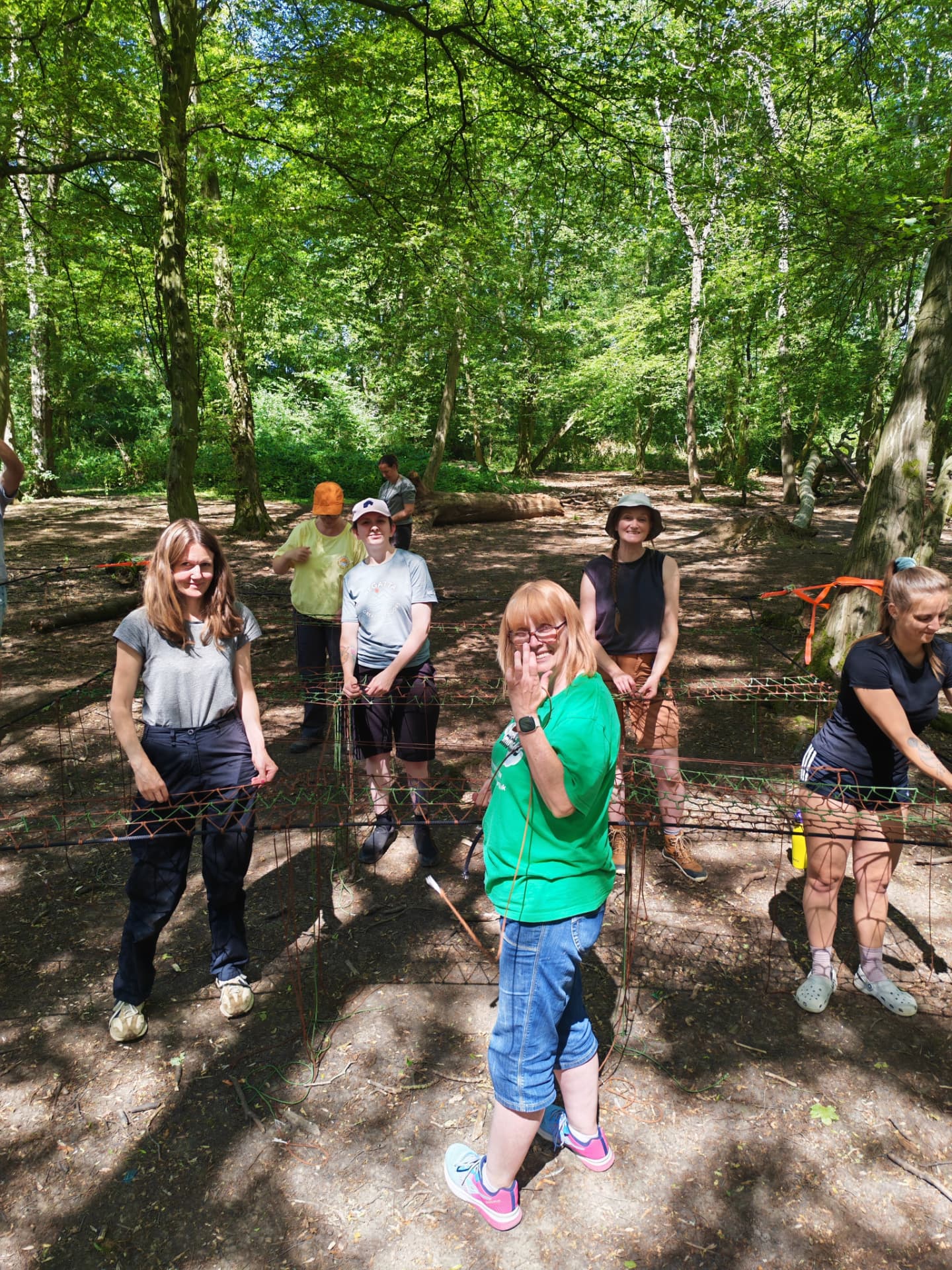 Team smiling while weaving net panels on frames in a sunlit woodland